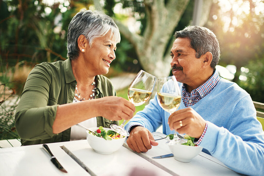 A senior couple toast with glasses of wine while enjoying a salad out on a patio table.