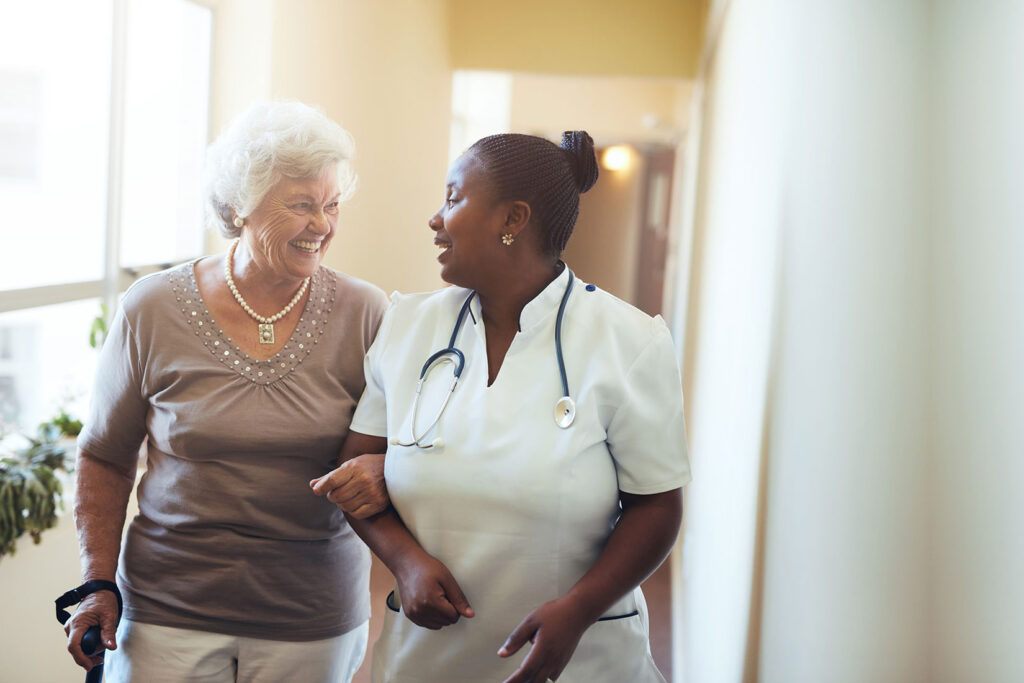 A senior woman walks with assistance from a nurse.
