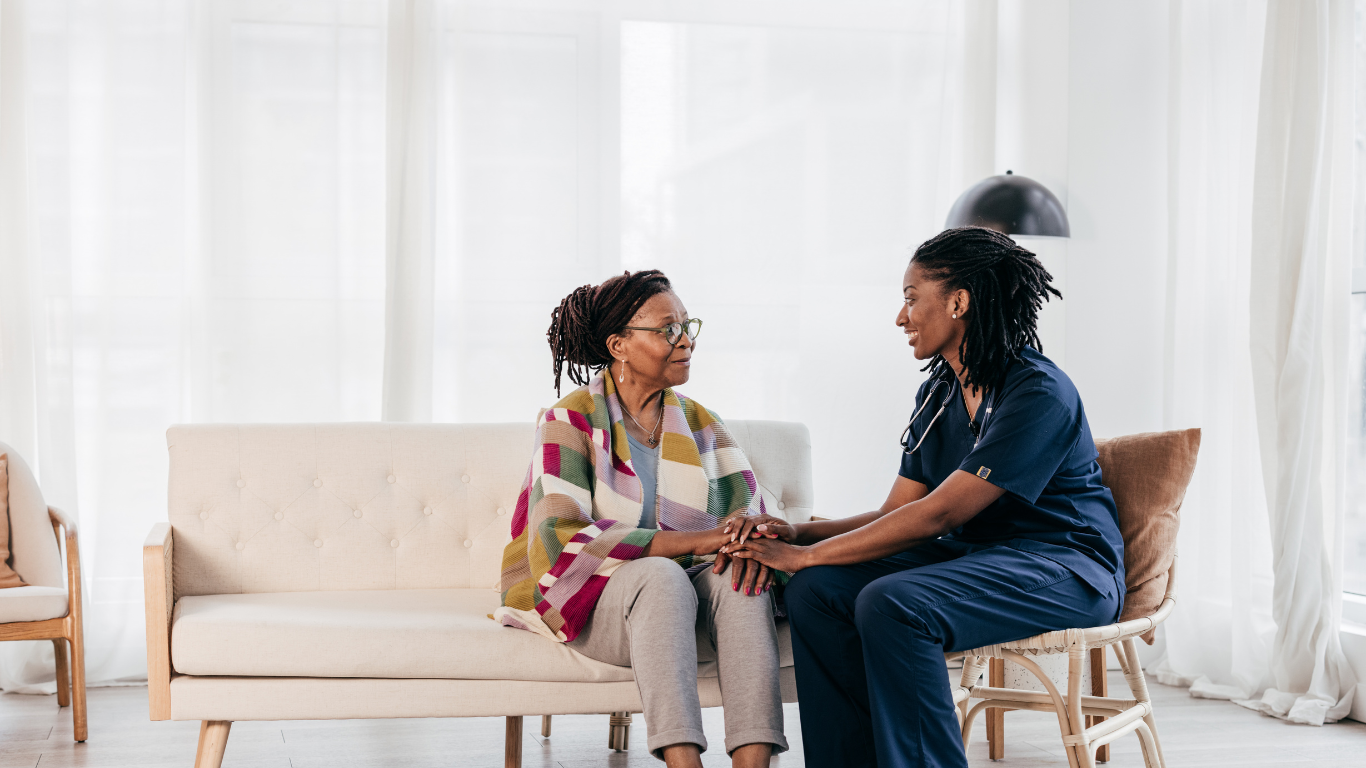 A health care worker sits next to a senior woman and holds her hands while talking to her