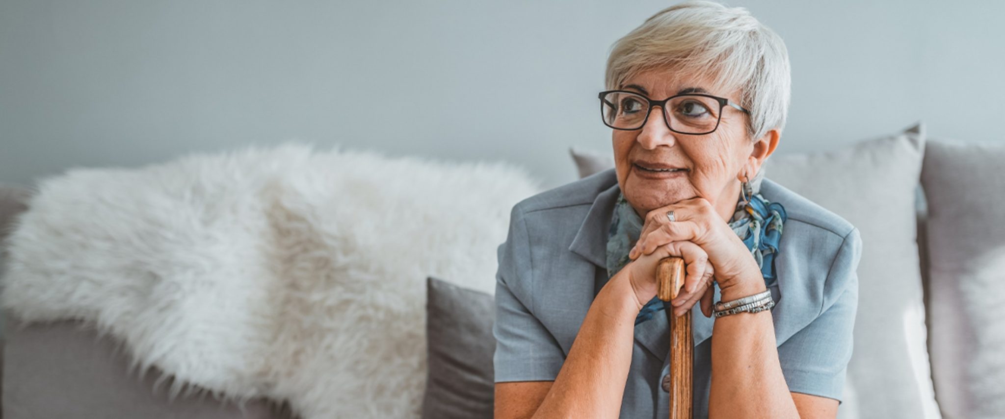 A senior woman sits on a couch and leans forward on her walking cane