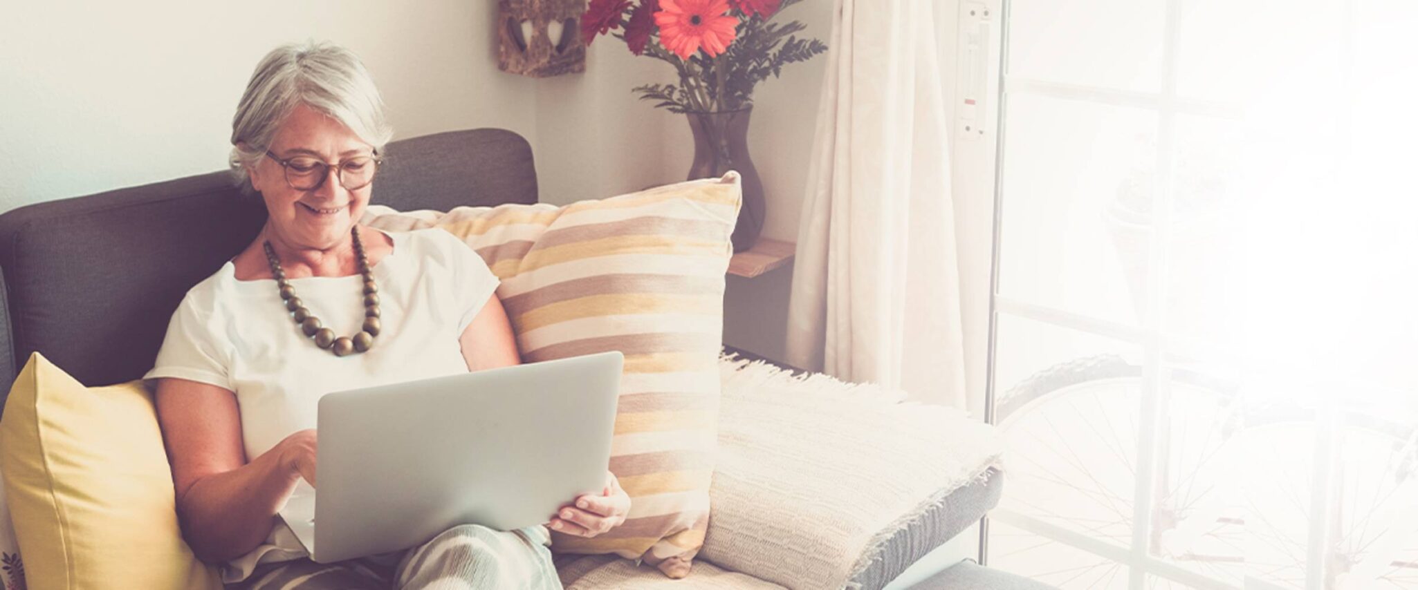 A senior woman sits comfortably on a couch in a sunny room looking at a laptop