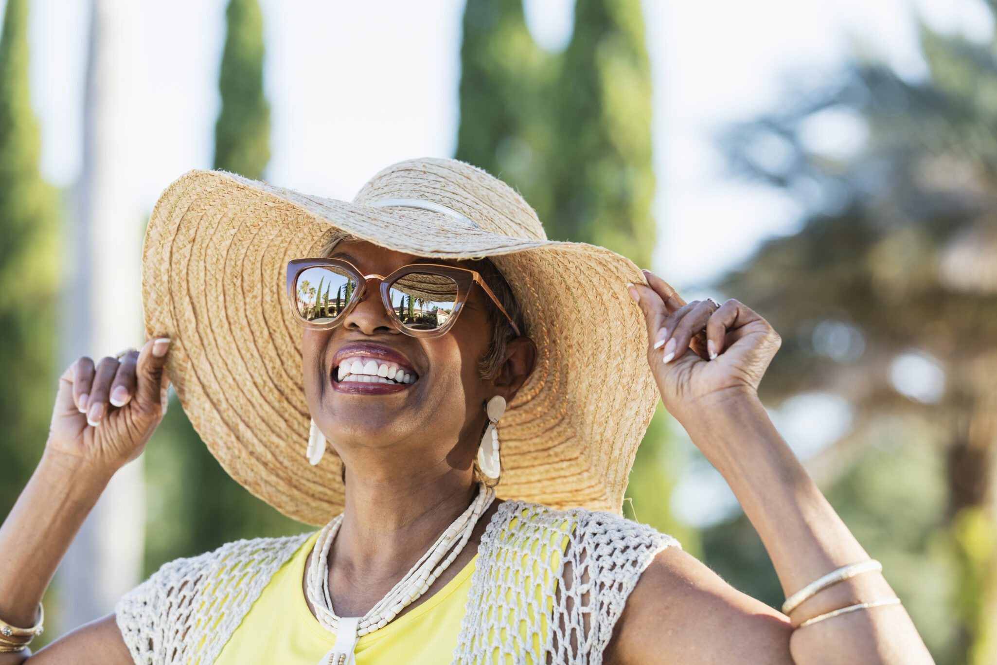 A women smiling widely while wearing sunglasses and touching the brim of her sun hat
