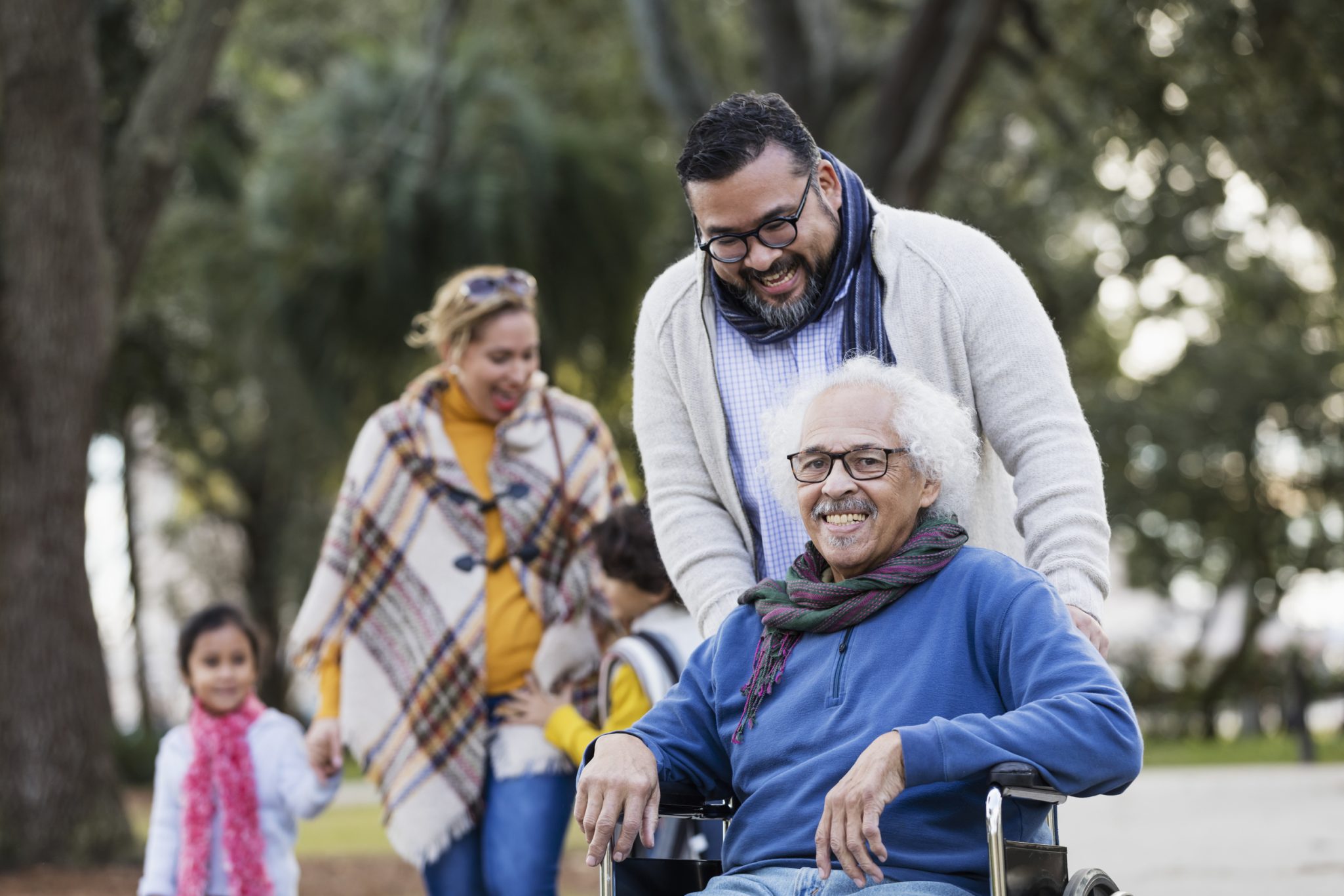 Smiling older man in a wheelchair being pushed by a family member outdoors, with other family members walking behind